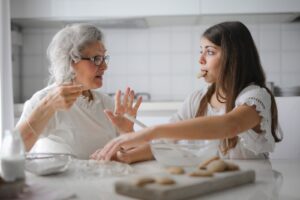 Calm senior woman and teenage girl in casual clothes looking at each other and talking while eating cookies and cooking pastry in contemporary kitchen at home